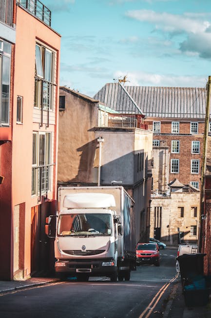 A narrow urban street scene in Bethnal Green during daytime, showing parked cars along the right side and a white delivery van from Man With a Van Bethnal Green positioned to load or unload furniture.