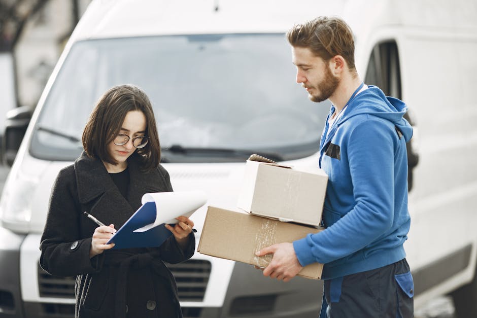 A young man with a serious expression, wearing a beige knit hat, a green padded vest over a dark blue jacket with white piping, and black gloves, is seated on the edge of an open van's loading area. B
