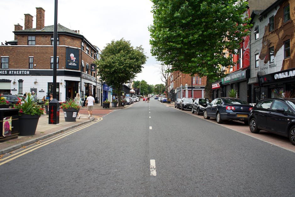 A wide residential street in Bethnal Green during daytime, with parked cars lining both sides of the road. The left pavement features potted plants with flowering arrangements and a few pedestrians walking, including a person carrying a box. At the end of the street, there are trees and more buildings in the distance. To the right, shopfronts and commercial buildings with signage are visible, while on the left, a corner building displays the logo of a pub or restaurant. A van belonging to Man With a Van Bethnal Green is shown in the process of loading furniture and boxes into its rear, part of a home relocation or furniture transport operation. The scene captures the organized parking, the urban environment, and the activity associated with packing and moving services, with natural lighting and clear visibility of the street layout, vehicles, and surroundings.