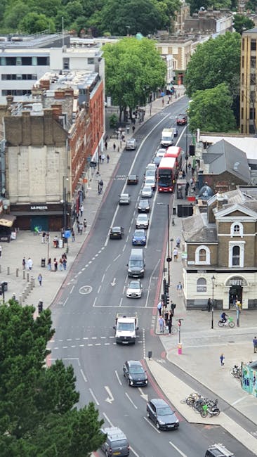 A high-angle view of Mile End Road in Bethnal Green, showing a busy urban street scene during daytime. The pavement is lined with pedestrians walking along the sidewalks, with some waiting at crossing