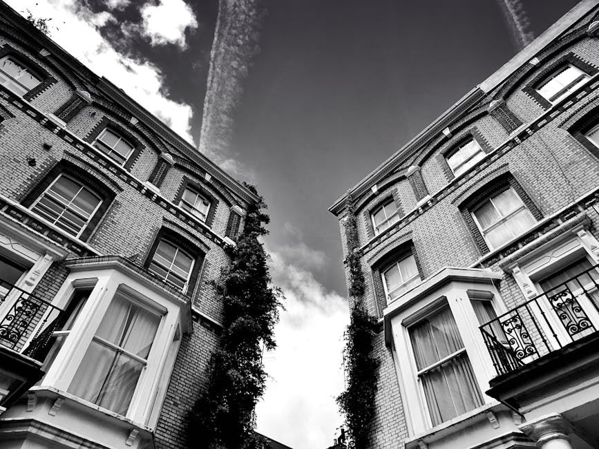 Black-and-white photograph of two Victorian-style brick residential buildings with multiple levels and bay windows, separated by a narrow alleyway. The buildings feature decorative cornices, wrought iron balcony railings, and large sash windows with curtains. Ivy or climbing plants ascend the sides of both structures, adding greenery to the façades. The sky above shows a mix of clouds and contrails, with sunlight casting subtle shadows on the buildings. The image captures the exterior view from a low angle, emphasizing the height of the buildings and the confined urban space typical of Bethnal Green. This scene relates to house removals and furniture transport in tight city environments, illustrating the challenges of home relocation and the importance of careful packing and moving logistics, as handled by Man With a Van Bethnal Green.