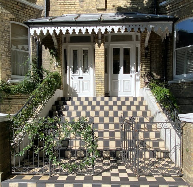 Exterior view of a Victorian residential building featuring a tiled staircase leading up to two white front doors with glass panels, situated beneath a decorative metal awning. The stairs are paved with black and beige checkered tiles, extending onto the pavement area. Black wrought iron railings with ornamental details run along each side of the staircase, with an additional small gate at the bottom, partially open. To the left, a leafy green shrub climbs the railing, casting dappled shadows on the tiled steps. The building’s facade is made of yellow brick, flanked by neighboring structures with similar brickwork and windows. The scene is illuminated by natural daylight, highlighting the textures of the brick, metalwork, and foliage, and capturing a typical urban home relocation environment involving furniture transport and packing preparations by Man With a Van Bethnal Green.