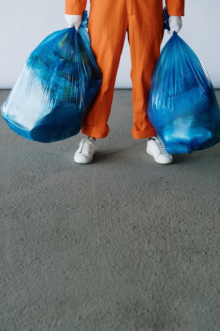 A large collection of mixed household waste, including paper, cardboard boxes, plastic bags, and larger items such as a damaged chair and packaging materials, piled next to multiple waste bins of vary